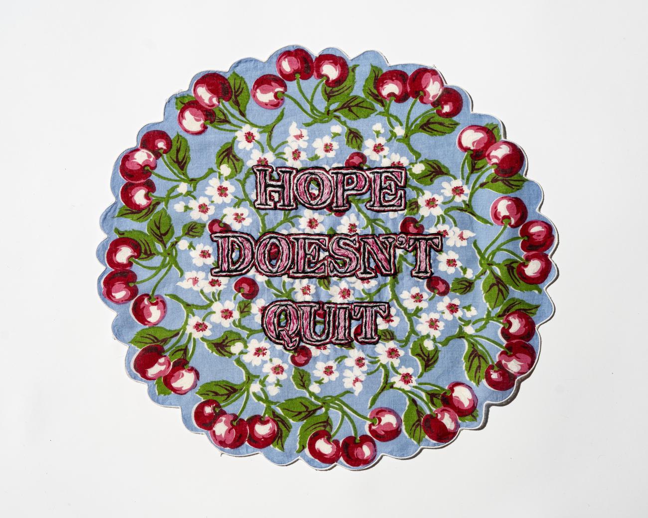 The words &quot;Hope doesn't quit&quot; is stiched onto a blue, round textile with flowers in the centre and cherries around the border.