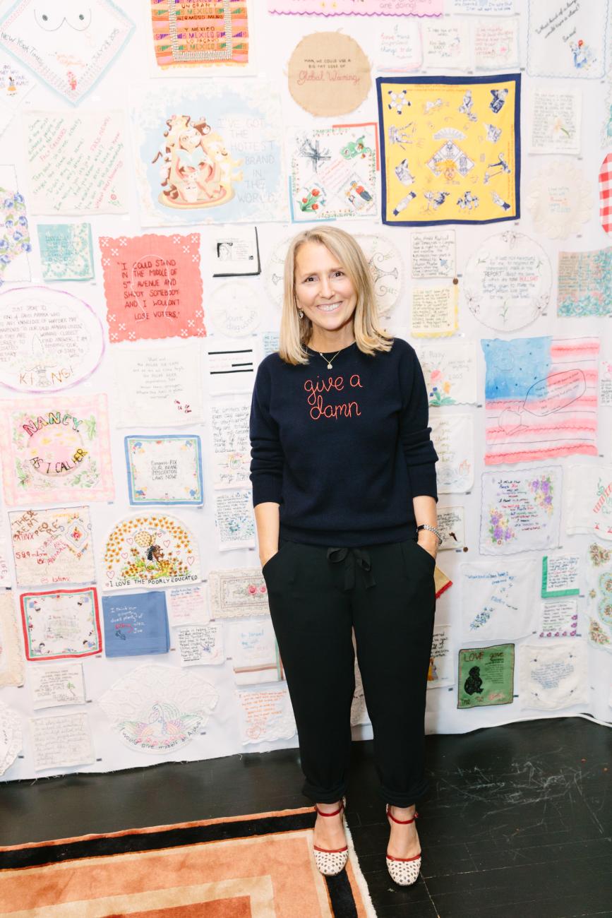 A photo of textile artist and activist Diana Weymar in front of a Gallery of works from Tiny Pricks Project. Her shirt has &quot;give a damn&quot; stitched on it.