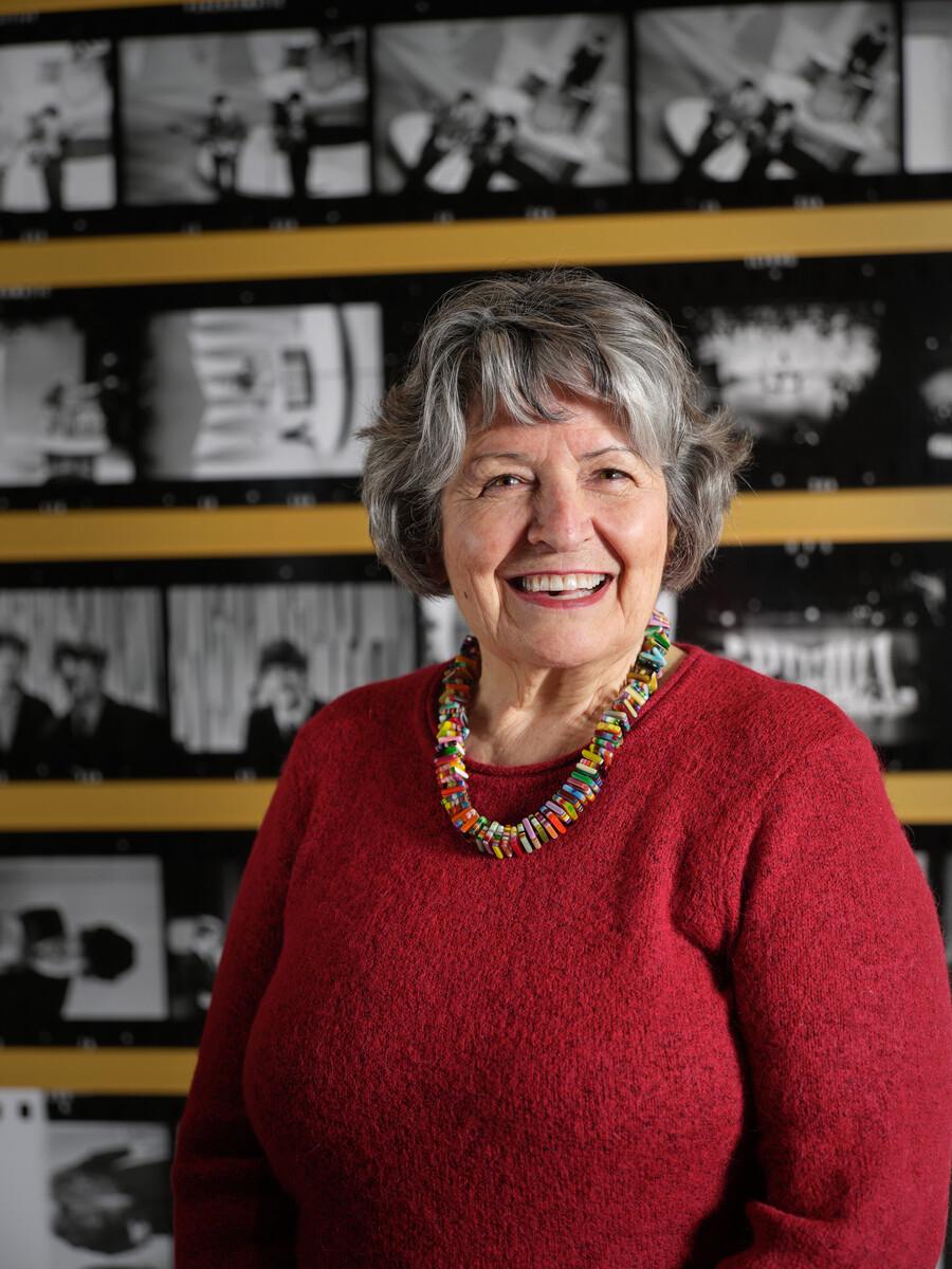 A headshot photograph of Trudy Medcalf. She smiles in front of a yellow wall featuring film strips of The Beatles in a red sweater.