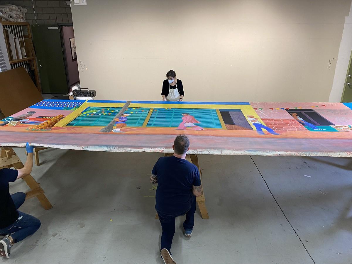 Studio assistants and David Hockney stand around the canvas as it lays flat on a table