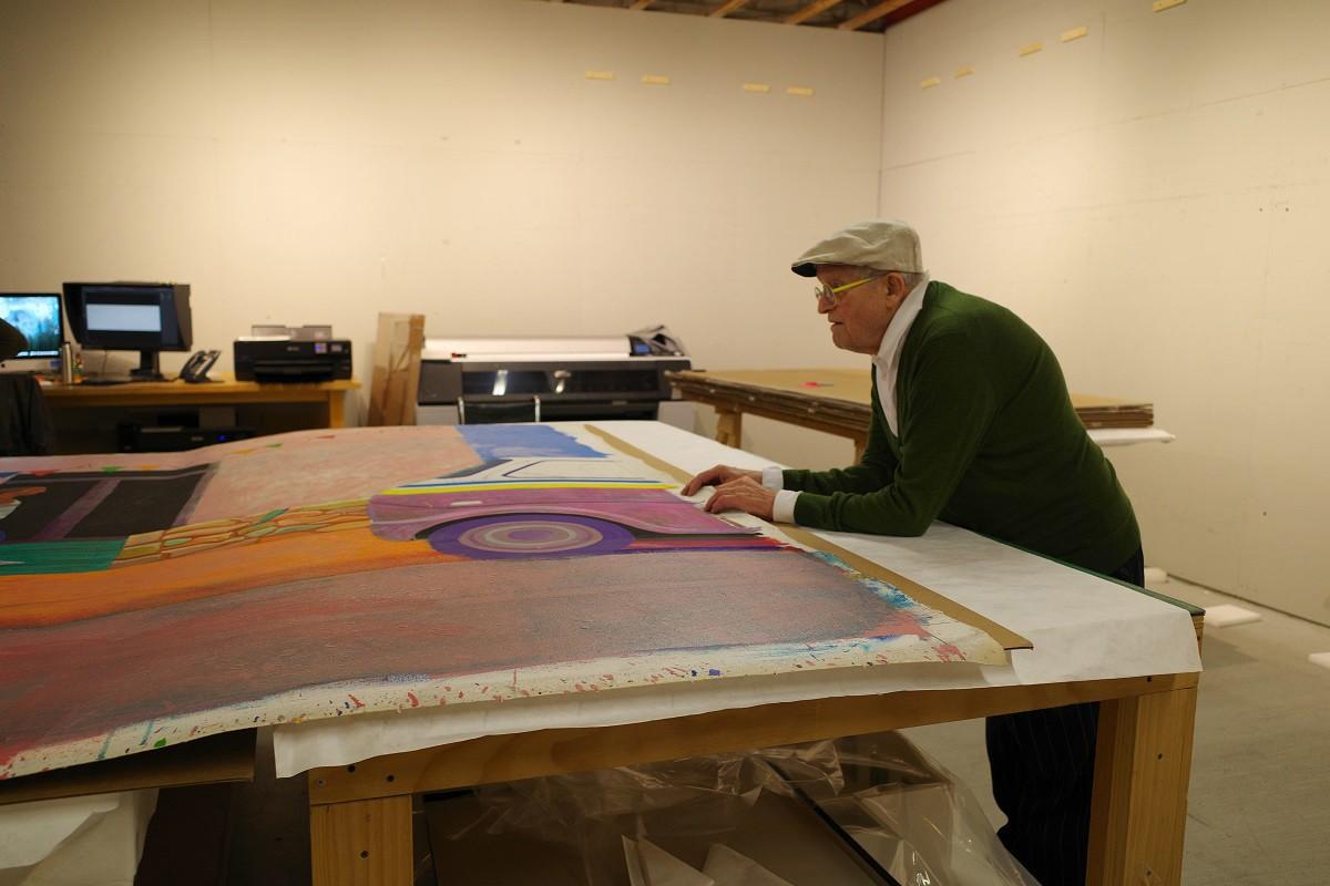 Studio assistants and David Hockney stand around the canvas as it lays flat on a table