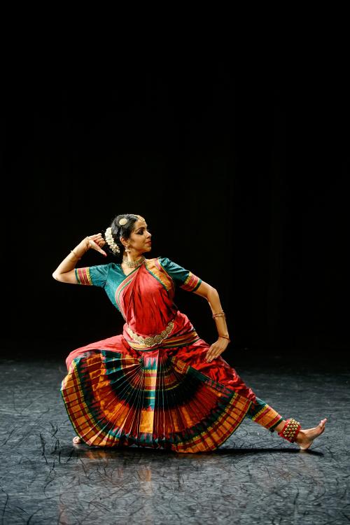 A photo of dancer Shilpa Shankar dancing on stage. Their left leg is outstretched with a flexed foot and their right hand is rested on the back of their head as they look to their left. 