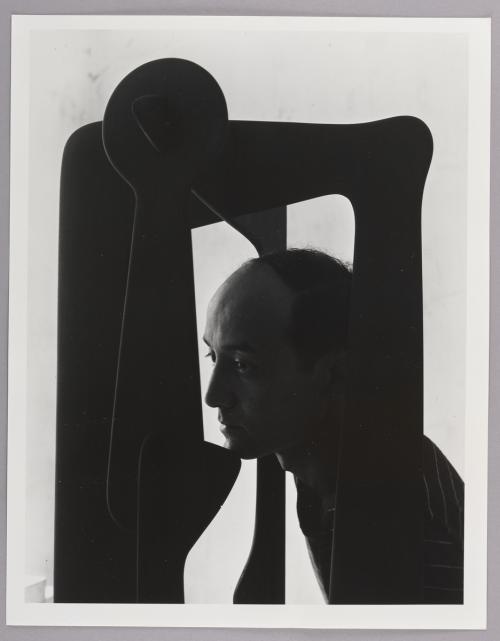 A black-and-white portrait of sculptor Isamu Noguchi. A man faces towards the left with a neutral facial expression. He is photographed with a curvilinear sculpture.