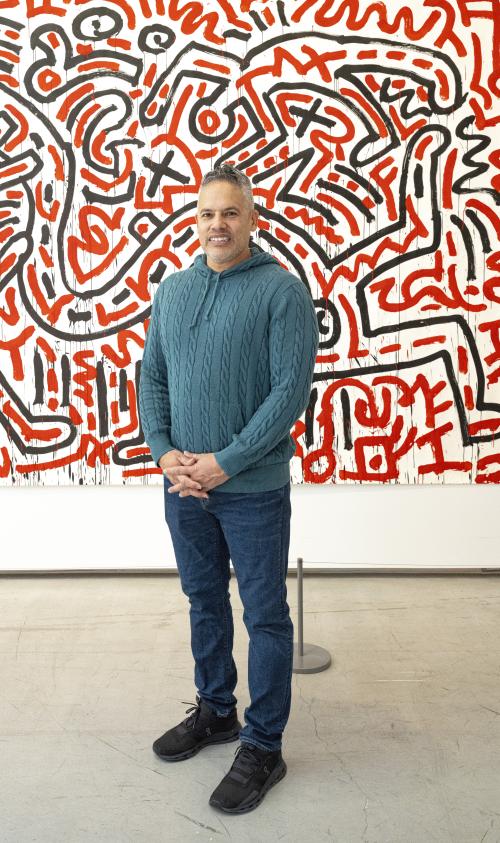 A person smiles while standing in front of a Keith Haring painting with black and red lines on a white background. 