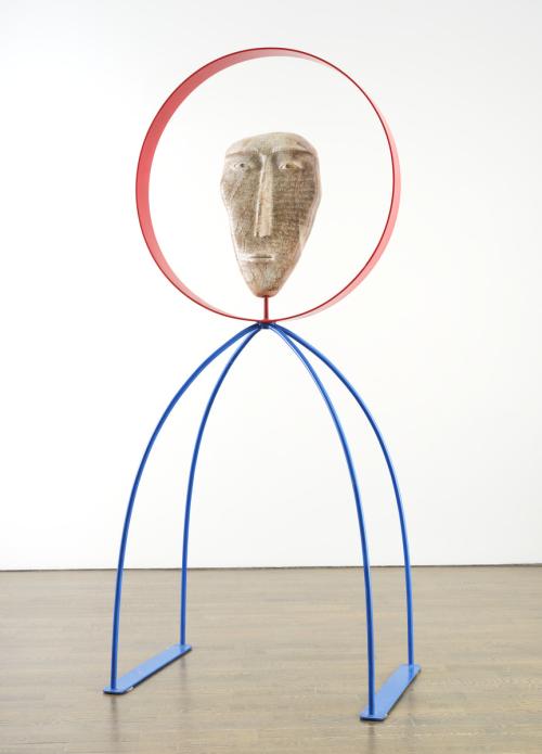 An image of David Ruben Piqtoukun's sculpture People of the Midnight Sun. It features a stone carved face with words carved into it, framed by a red metal circle. The statue stands on two metal blue legs that arch outwards. 